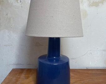 May include: A blue ceramic table lamp with a beige linen lampshade. The lamp has a rounded base and a cylindrical neck.