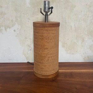May include: A brown cylindrical lamp base with a silver lamp socket and a black cord. The lamp base is made of woven material and has a wood top and bottom.