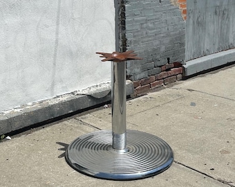 Könnte beinhalten: Ein Tischgestell aus Chrom mit einer kreisförmigen, gerippten Bodenplatte und einer zentralen zylindrischen Säule. Oben auf der Säule befindet sich eine flache, vierzackige Metallplatte mit rostbrauner Oberfläche zur Tischplattenmontage.