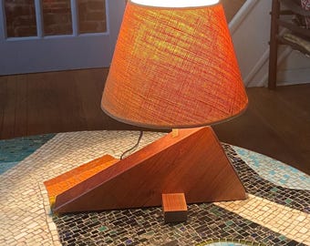 May include: A modern table lamp with a geometric wooden base and a conical orange fabric shade. The lamp is illuminated, casting a warm glow. The lamp sits on a mosaic table.