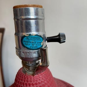 K&ouml;nnte beinhalten: Eine rote, strukturierte Lampenbasis mit einer silbernen Fassung und einem schwarzen Schalter. Die Fassung tr&auml;gt ein Etikett mit der Aufschrift "Underwriters Laboratories Inc. Portable Lamp Issue No. K-8349 E-61274"