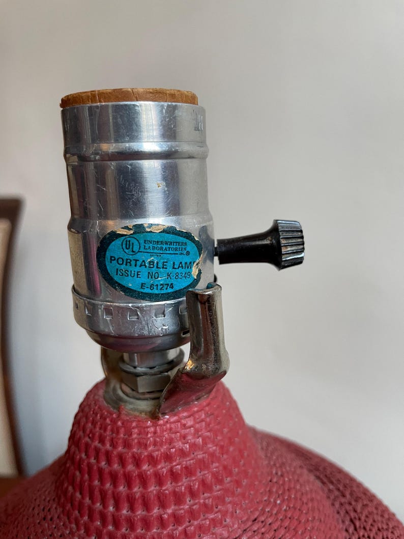 K&ouml;nnte beinhalten: Eine rote, strukturierte Lampenbasis mit einer silbernen Fassung und einem schwarzen Schalter. Die Fassung tr&auml;gt ein Etikett mit der Aufschrift "Underwriters Laboratories Inc. Portable Lamp Issue No. K-8349 E-61274"