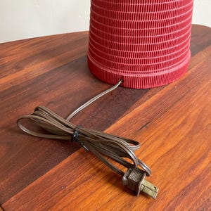 K&ouml;nnte beinhalten: Eine rote Keramik-Tischlampe mit einem strukturierten Sockel und einem braunen Stromkabel mit Stecker.