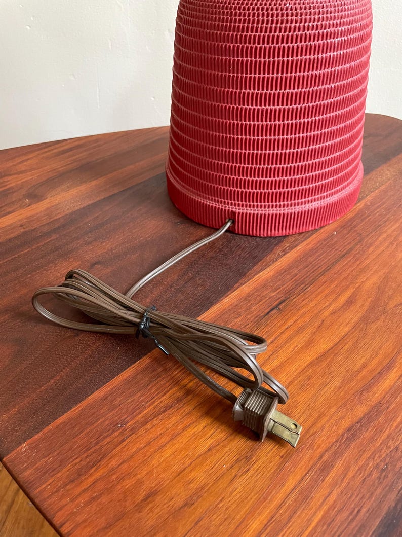 K&ouml;nnte beinhalten: Eine rote Keramik-Tischlampe mit einem strukturierten Sockel und einem braunen Stromkabel mit Stecker.