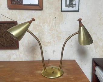 May include: A vintage brass desk lamp with two adjustable gooseneck arms and conical shades. The lamp features a round base and wooden accents on the top of each shade. A classic mid-century modern design.