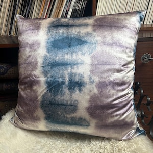 May include: A square decorative pillow with a tie-dye pattern in shades of blue, white, and purple. The pillow is made of a soft, velvety fabric. The pillow is resting on a white, fluffy surface.