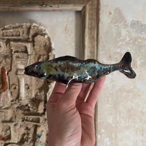 May include: A ceramic fish figurine with a blue, green, and brown glaze. The fish has a slightly open mouth and a tail that curves to the right.