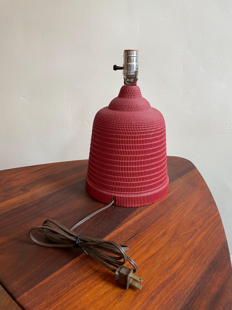 K&ouml;nnte beinhalten: Eine rote Keramik-Tischlampe mit einem strukturierten, gerippten Design. Die Lampe hat eine schlichte, klassische Form und ein braunes Kabel.