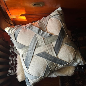 May include: A decorative square pillow with a woven design of gray and blue ribbons. The pillow is resting on a white, fluffy surface. The pillow is approximately 18 inches square.