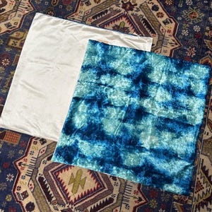 May include: Two decorative pillow covers. One is a solid off-white color, and the other features a tie-dye pattern in shades of blue and turquoise. The pillow covers are rectangular and made of a soft, textured fabric.
