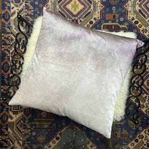 May include: A large, square, light purple velvet pillow rests on a white faux fur surface, set against a patterned rug. The pillow is the focal point, with a soft, textured appearance. The rug has a dark blue background with intricate patterns.