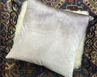 Könnte beinhalten: Ein großes, quadratisches, hellviolettes Samtkissen liegt auf einer weißen Kunstfelloberfläche, vor einem gemusterten Teppich. Das Kissen ist der Mittelpunkt, mit einem weichen, strukturierten Aussehen. Der Teppich hat einen dunkelblauen Hintergrund mit aufwendigen Mustern.