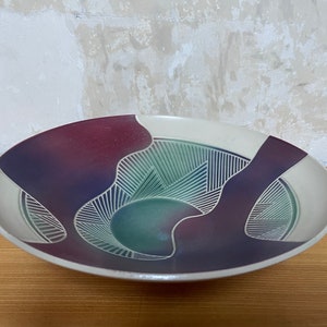 May include: A ceramic serving bowl with a unique design. The bowl features a cream-colored rim and a central design in shades of purple, green, and blue. The design includes geometric shapes and lines. The bowl is placed on a wooden surface.