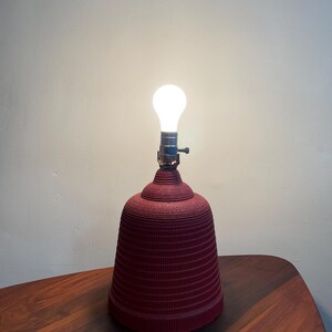 K&ouml;nnte beinhalten: Eine rote, wellpappenartige Lampe mit einer wei&szlig;en Gl&uuml;hbirne. Die Lampe steht auf einem Holztisch.