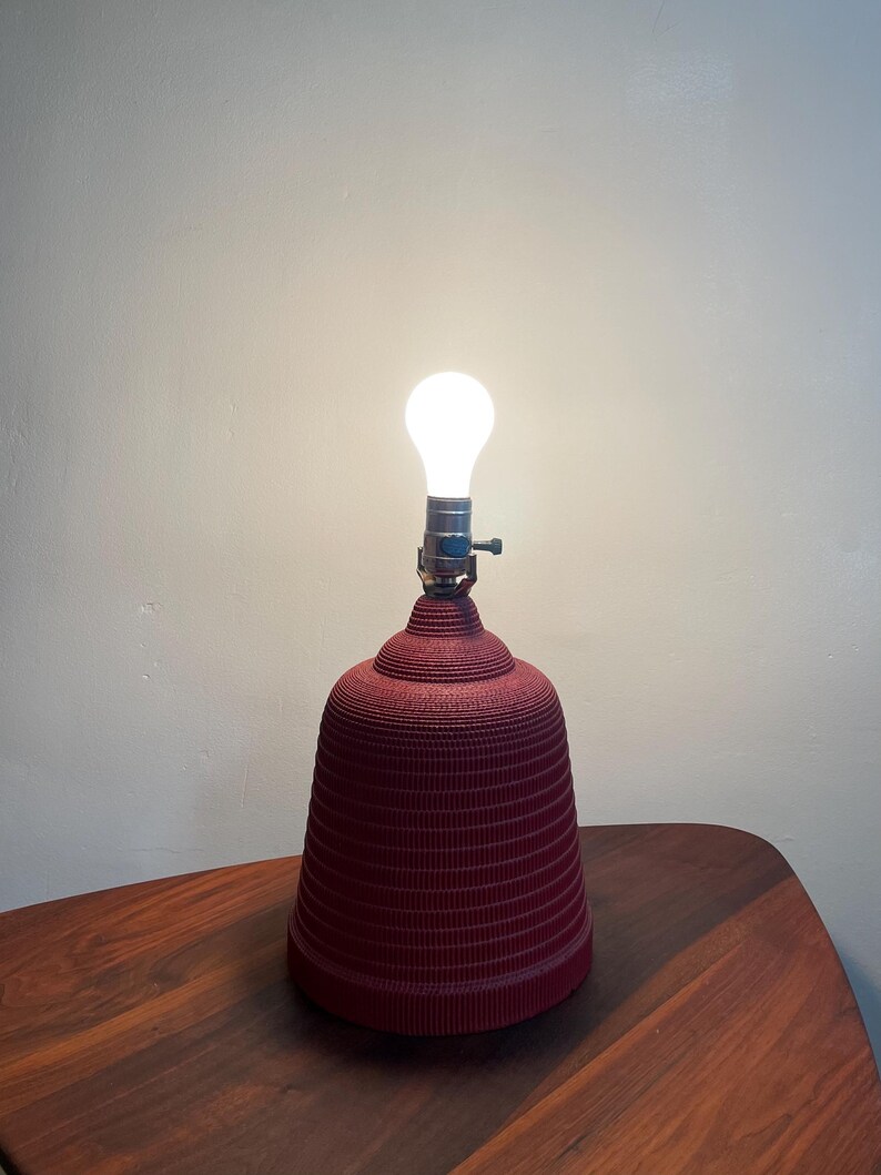 K&ouml;nnte beinhalten: Eine rote, wellpappenartige Lampe mit einer wei&szlig;en Gl&uuml;hbirne. Die Lampe steht auf einem Holztisch.