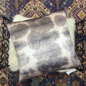 May include: A square decorative pillow with a gray and white pattern, resting on a white sheepskin rug. The pillow is placed on a black metal bed frame, with a colorful patterned rug in the background.