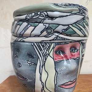 May include: A ceramic vase with a face design. The vase is white with green, blue, and pink accents. The face is made up of different patterns and lines.