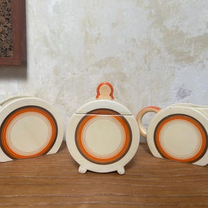 May include: A vintage cream-colored ceramic tea set with orange and brown concentric circle designs. The set includes a sugar bowl with a lid, a creamer, and a small dish. The set is displayed on a wooden surface.