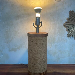 May include: A cylindrical table lamp with a woven paper shade. The lamp has a silver metal base and a light bulb is turned on.