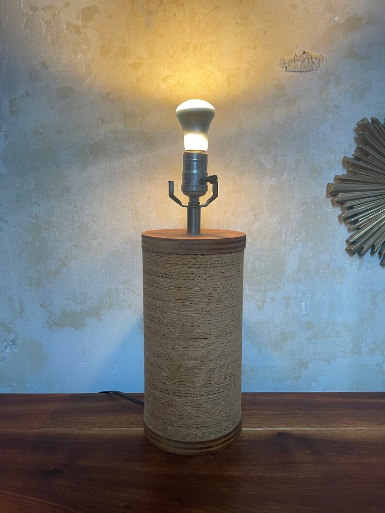 May include: A cylindrical table lamp with a woven paper shade. The lamp has a silver metal base and a light bulb is turned on.