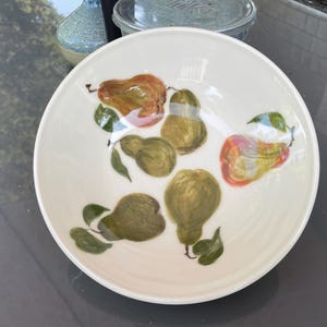Könnte beinhalten: Eine weiße Keramikschüssel mit handbemaltem Design aus grünen und braunen Birnen. Die Birnen sind in einer Gruppe angeordnet, wobei sich einige überlappen. Die Schüssel hat eine leicht gewellte Oberfläche.