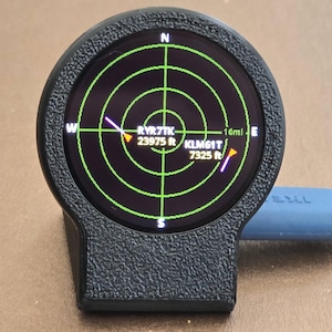 May include: A black circular electronic device with a textured frame displaying a green radar screen interface. The screen shows flight data including RYR7TK at 23975 ft and KLM61T at 7325 ft with cardinal directions. A blue cable is plugged into the side.