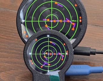 May include: Two circular black radar displays with green grid lines and orange directional markers. The screens show a 25km range indicator and cardinal directions N, W, and S. A blue USB cable is connected to the smaller unit, which features a small green square sticker on its base.