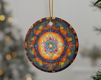 Op de afbeelding: Een ronde keramische hanger met een levendig, symmetrisch mandala-ontwerp met een centraal zonnepatroon in tinten oranje, geel, blauw en paars. De hanger hangt aan een dun gouden koordje en toont ingewikkelde, kleurrijke bloemachtige blaadjes.