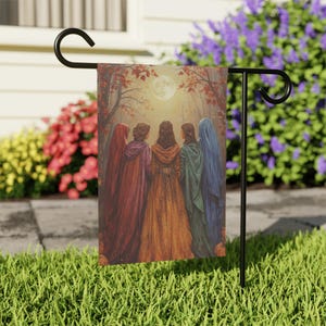 Puede incluir: Una bandera de jardín rectangular colgada en un soporte de metal negro, con una ilustración de cinco figuras con capas con capucha en rojo, morado, naranja, verde y azul, de pie en un bosque bajo una luna llena con hojas de otoño y calabazas.
