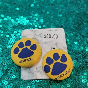 Può includere: Un paio di orecchini in tessuto giallo con un'impronta di zampa blu e la parola "Cougar" su ogni orecchino. Gli orecchini sono su una superficie di marmo bianco con un'etichetta del prezzo che dice 10,00 €.
