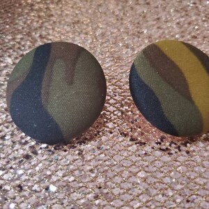 May include: Two round stud earrings with a camouflage pattern. The earrings feature a fabric-covered design in shades of green, brown, and black. The earrings are circular and have a button-like appearance.