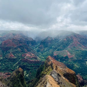 May include: A view from a cliff overlooking a deep canyon with lush green vegetation and red rock formations. The sky is cloudy and the canyon is shrouded in mist.