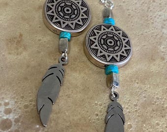 May include: Silver earrings with turquoise beads and a silver feather charm. The earrings feature a circular pendant with a starburst design.