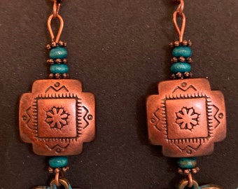 May include: A pair of copper earrings with turquoise accents. The earrings feature a square charm with a floral design and a heart-shaped charm.