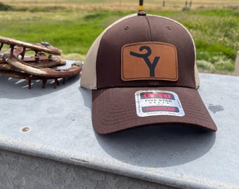 May include: A brown trucker hat with a tan mesh back and a tan leather patch on the front featuring a black stylized Y logo. The hat has a curved brim with a sticker that reads OTTO FIVE STAR TRUCKER CLASSIC MESH BACK.