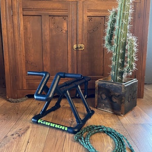 May include: A black metal bike stand with the word "Kannon" printed on it. The stand is on a wooden floor next to a green rope and a potted cactus.