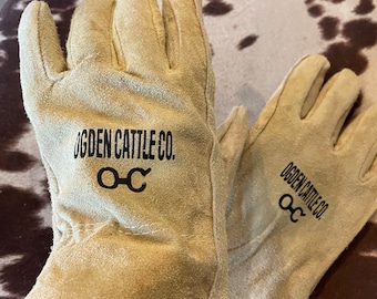 May include: A pair of tan leather work gloves with a green trim. The gloves have the text "Ogden Cattle Co. OCC" printed on them.