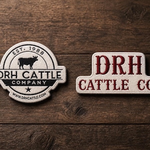 May include: Two embroidered patches on a wooden surface. One is circular with a black cow silhouette, the text "DRH CATTLE COMPANY", and "EST. 1988". The other is rectangular with "DRH CATTLE CO." in burgundy.