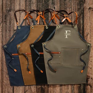May include: Four canvas aprons with adjustable leather straps and a large pocket. The aprons are in navy blue, brown, black, and olive green. The olive green apron has the letter "F" and the word "Flores" embroidered on it.