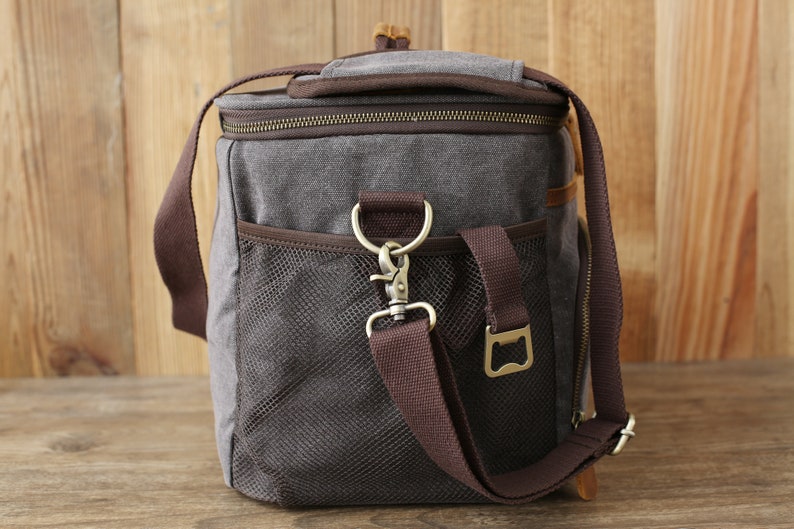 Puede incluir: Una bolsa isot&eacute;rmica de lona gris con una correa de cuero marr&oacute;n y un abridor de botellas. La bolsa tiene un bolsillo de malla en el lateral y un cierre de cremallera.