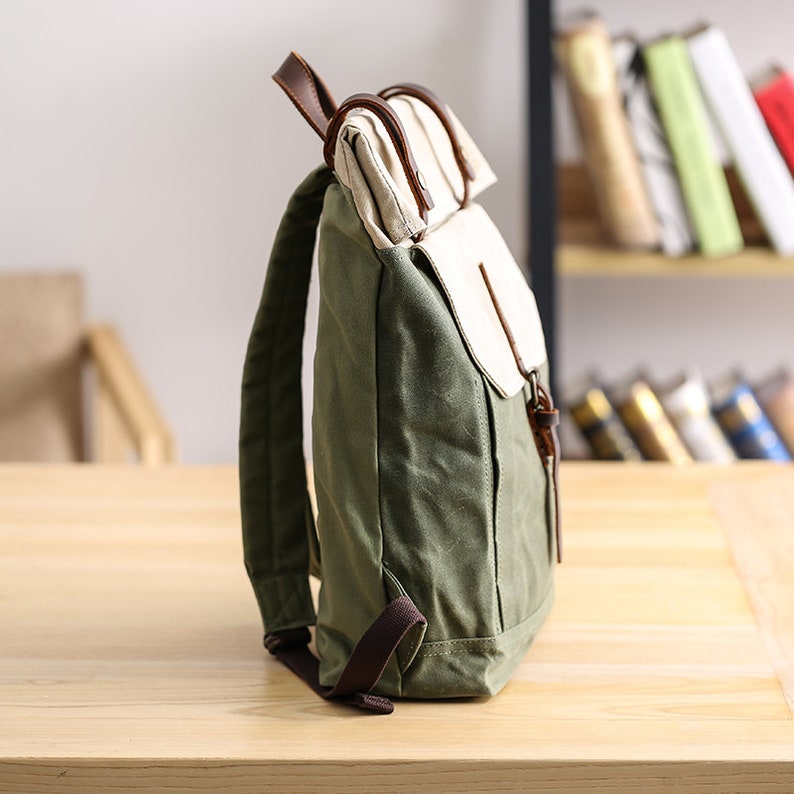 May include: A green canvas backpack with a brown leather flap and straps. The backpack has a white canvas pocket on the front.
