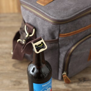 Puede incluir: Una bolsa de enfriador de lona gris con detalles de cuero marr&oacute;n y cierre de cremallera. Una botella marr&oacute;n con una etiqueta azul est&aacute; unida a la bolsa con un abridor de botellas incorporado.