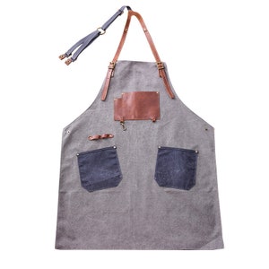 May include: Gray canvas apron with brown leather accents, including straps and a chest patch. Features two dark blue denim pockets and a small metal hook. Adjustable neck strap and waist ties.