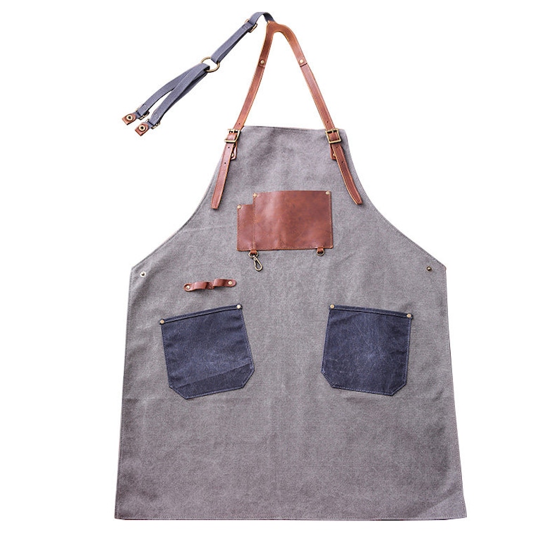 May include: Gray canvas apron with brown leather accents, including straps and a chest patch. Features two dark blue denim pockets and a small metal hook. Adjustable neck strap and waist ties.