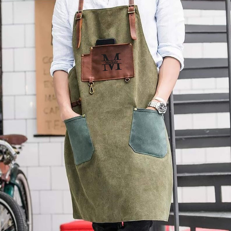 May include: An olive green apron with brown leather straps and accents. The apron has two front pockets with green suede-like fabric and a brown leather pocket with the initials "M IVI" printed on it. A phone is in the top pocket.