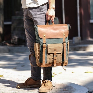 May include: A green and brown canvas backpack with leather accents and buckles. The backpack has a large main compartment and a smaller front pocket.