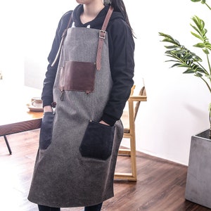 May include: A gray apron with brown leather accents, including a chest pocket and shoulder straps. The apron has two large black pockets and a metal clasp. The apron is designed for kitchen or workshop use.