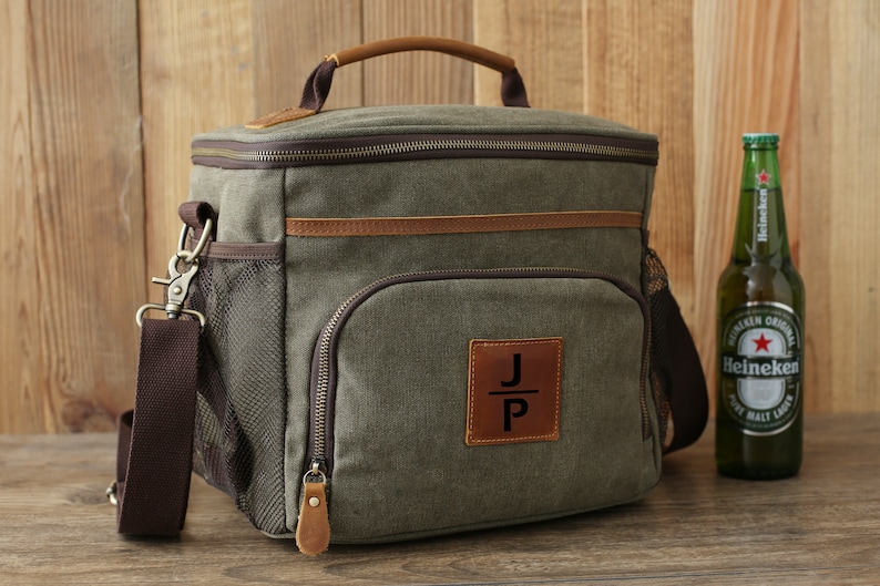 Puede incluir: Una bolsa de enfriador de lona verde con detalles de cuero marr&oacute;n y cierre de cremallera. La bolsa tiene una correa para el hombro y un bolsillo frontal con un parche de cuero que dice "J+P".