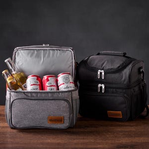 Puede incluir: Dos bolsas de almuerzo aisladas con cierres de cremallera. La bolsa gris está abierta y contiene cuatro latas de cerveza y dos botellas de cerveza. La bolsa negra está cerrada. Ambas bolsas tienen un parche de cuero con el nombre de la marca.