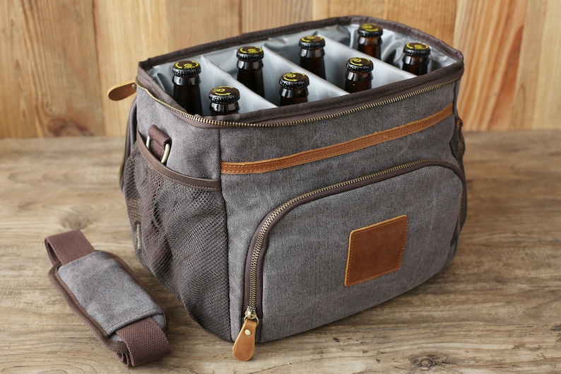 Puede incluir: Una bolsa de enfriador de lona gris con ribete de cuero marr&oacute;n y un bolsillo con cremallera. La bolsa de enfriador tiene una correa para el hombro y est&aacute; llena de seis botellas de vidrio marr&oacute;n.